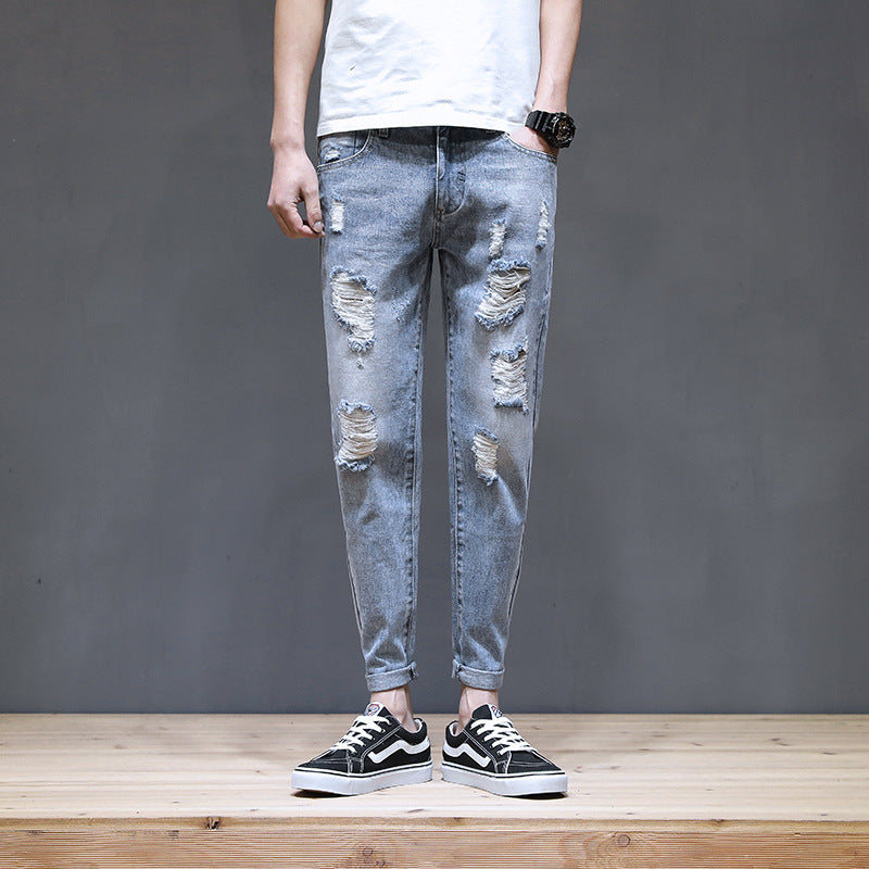 Person wearing distressed light blue jeans and black sneakers on a wooden floor with a gray wall background