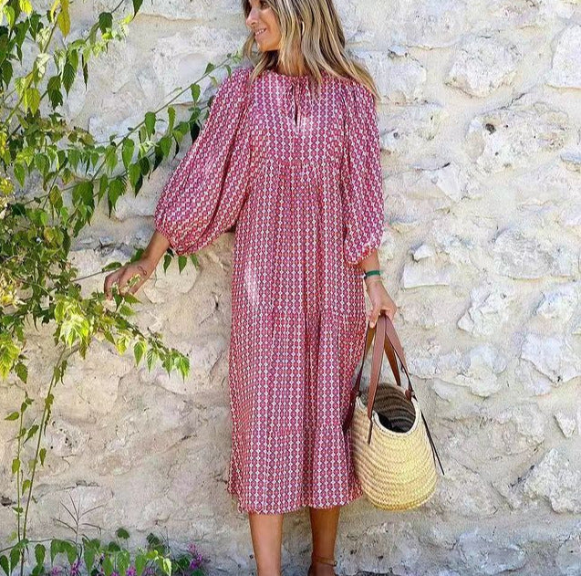 Woman in a pink patterned dress holding a straw bag against a stone wall.